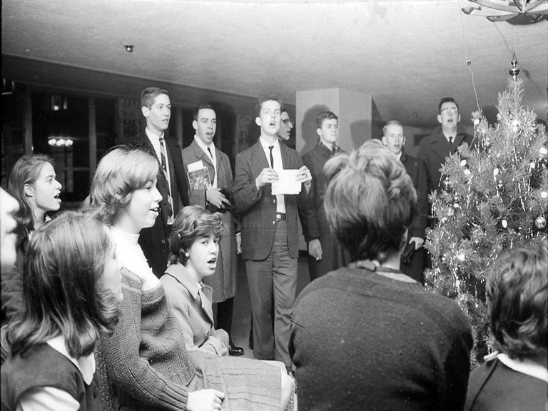 A black and white photograph features a group of seven young men standing and about five young women sitting, one with her back to the camera, in front of a Christmas tree, singing carols.
